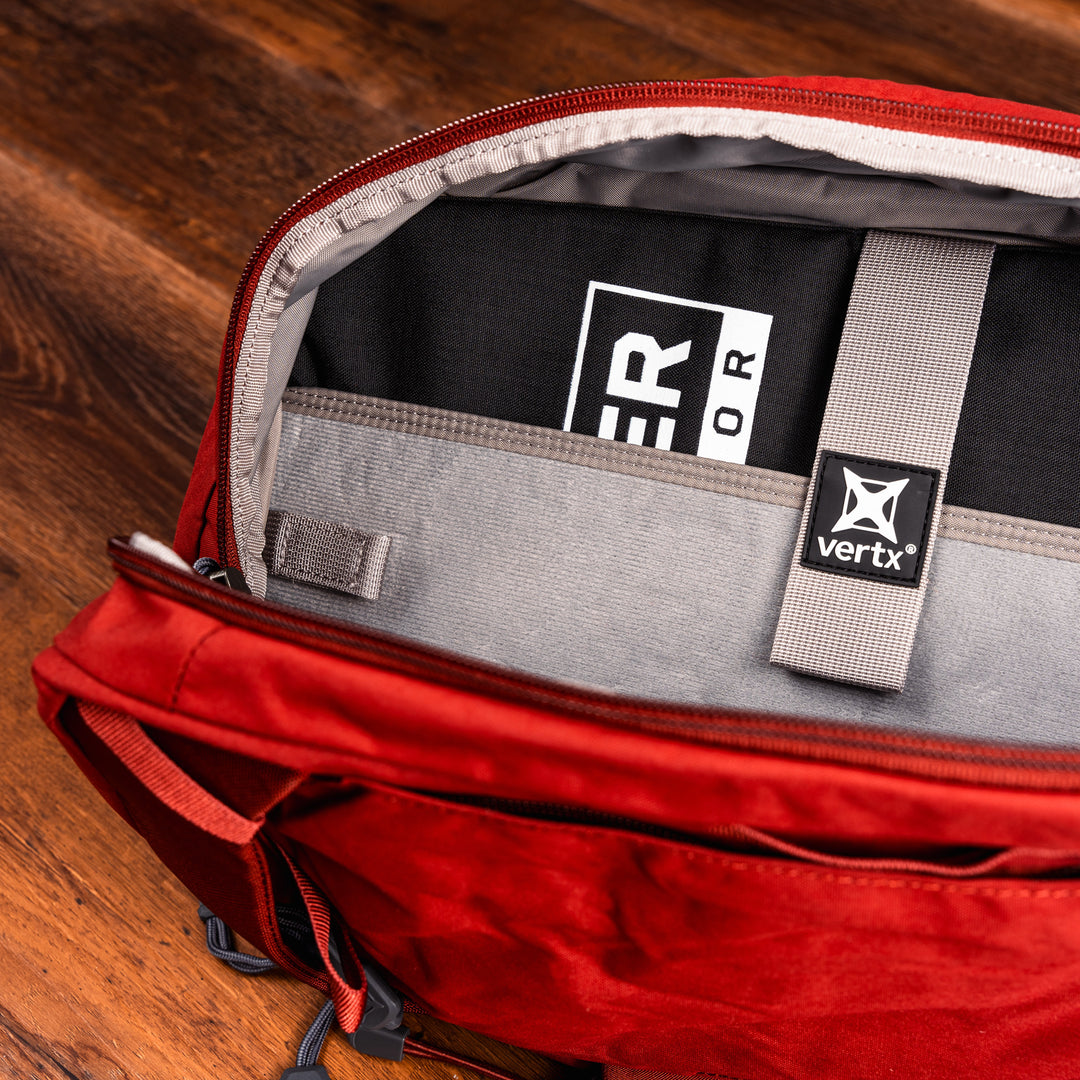 A red backpack is open, revealing a black and white labeled item inside. The interior has a gray compartment with a "vertx" label. The bag is placed on a wooden surface.