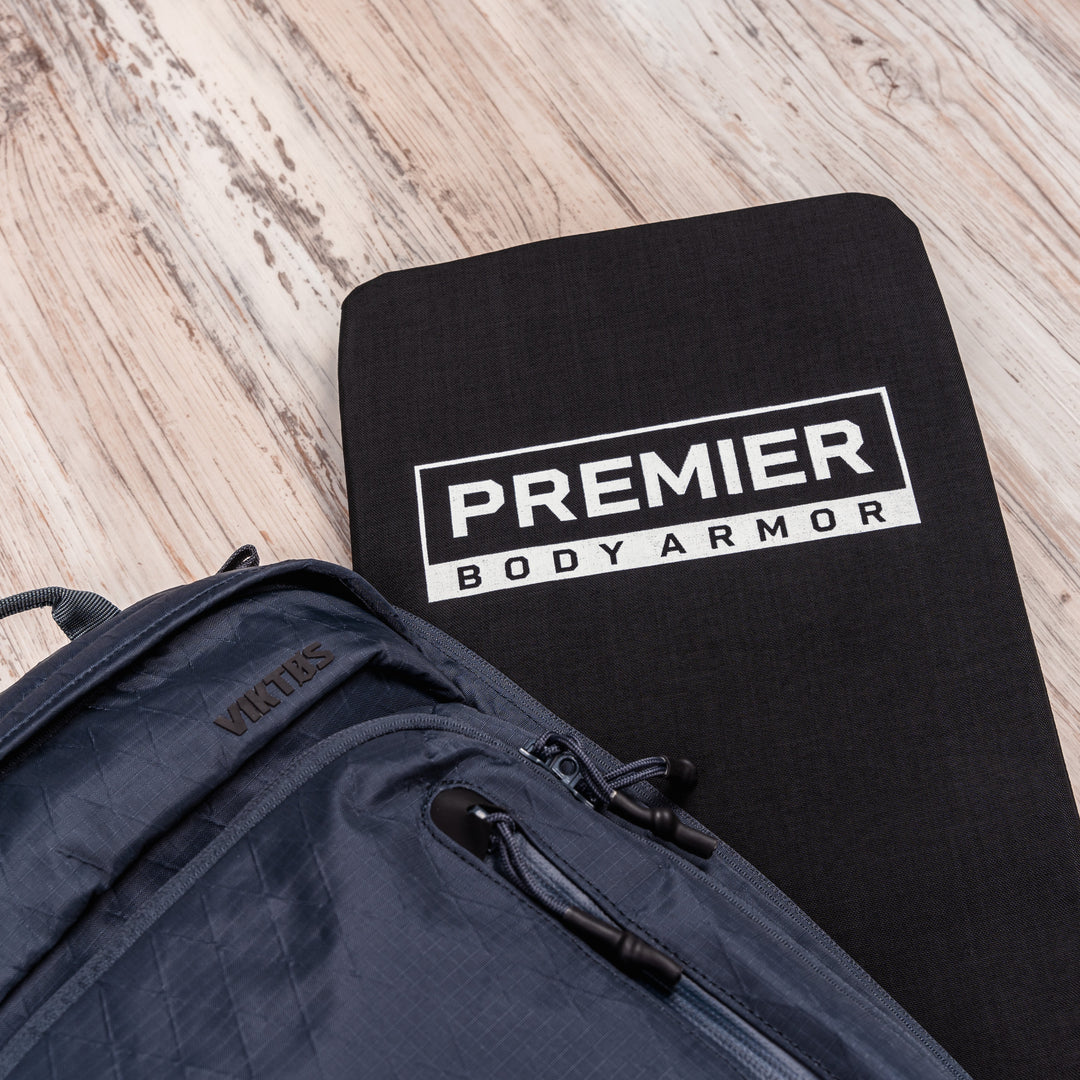 A black protective panel labeled "Premier Body Armor" is placed on a light wood surface next to a dark gray backpack branded with "VIKTOS".