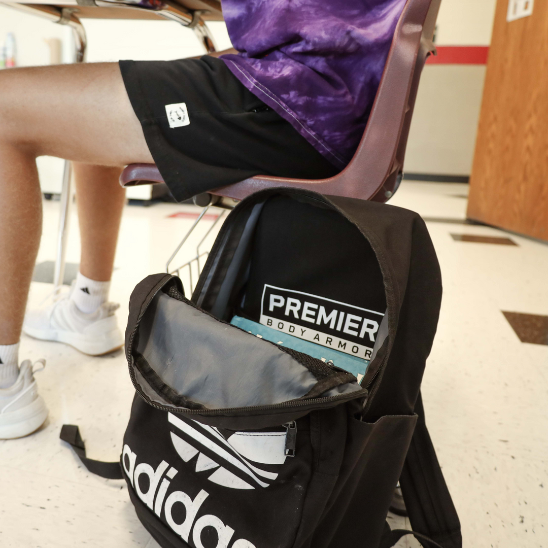 A person wearing a purple shirt and black shorts sits on a chair. A black Adidas backpack with a Premier Body Armor panel inside is on the floor nearby.