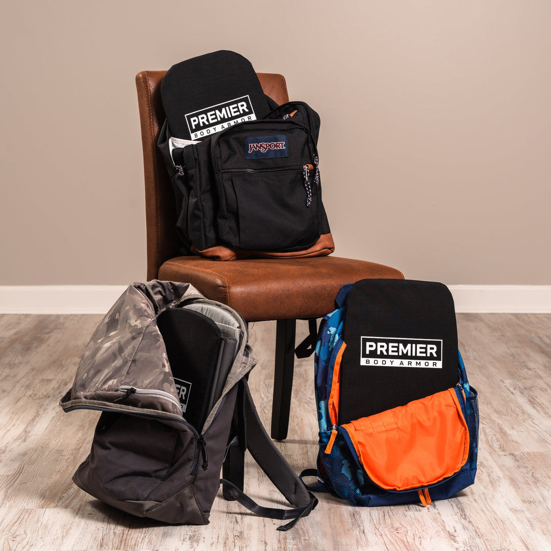 Three backpacks, each with a black Premier Body Armor insert visible, are displayed; one black backpack sits on a brown chair, while a gray and a blue/orange backpack rest on the wooden floor.