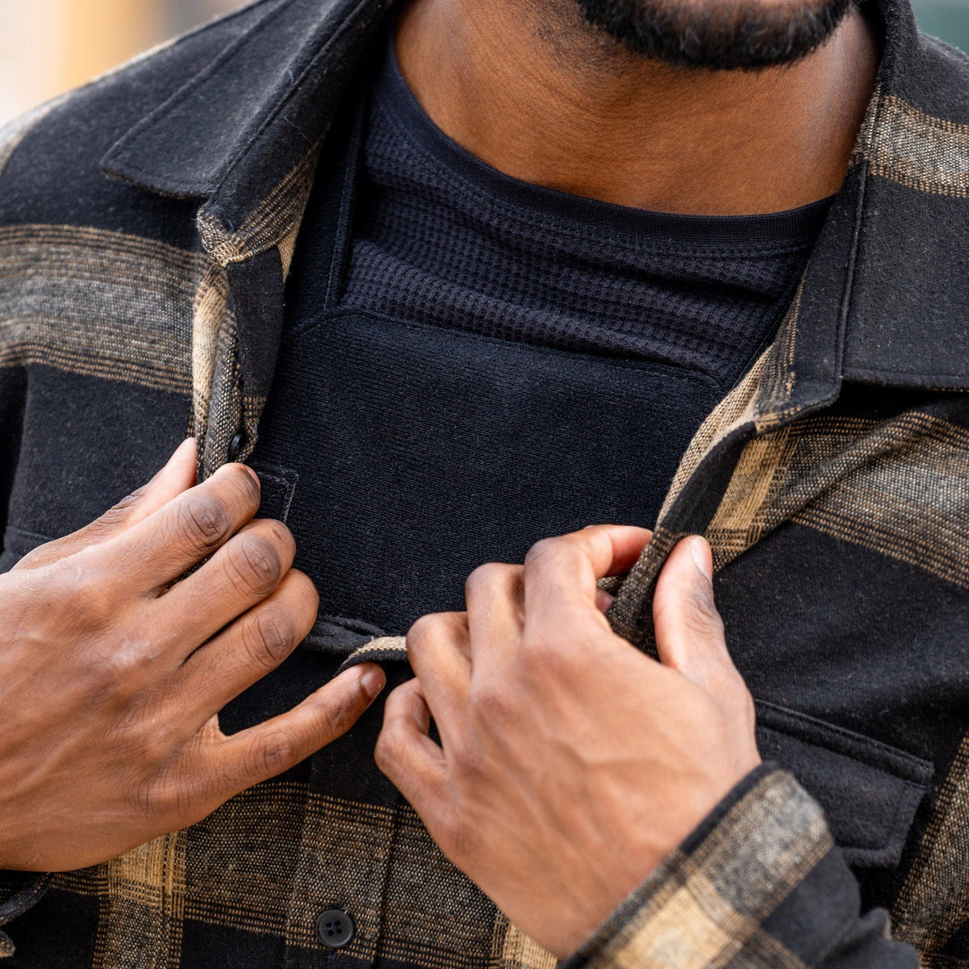 A person in a black and brown plaid jacket over a black shirt adjusts their jacket, partially revealing the Premier Body Armor Discreet Plate Carrier underneath, with their hands near the chest. The background is blurred.