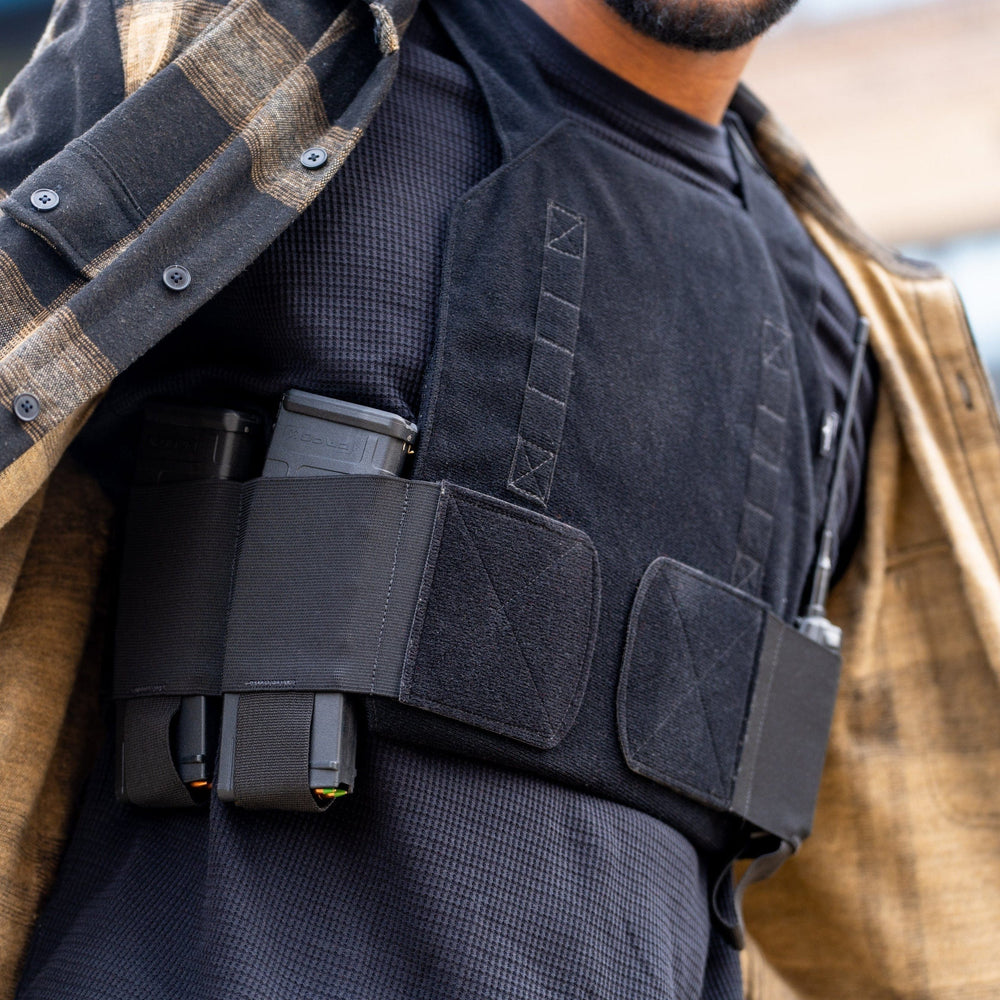 A person wears a black tactical vest under an open brown and black plaid shirt. The Premier Body Armor Discreet Plate Carrier, lightweight and quiet, holds two magazines without fully showing its design.