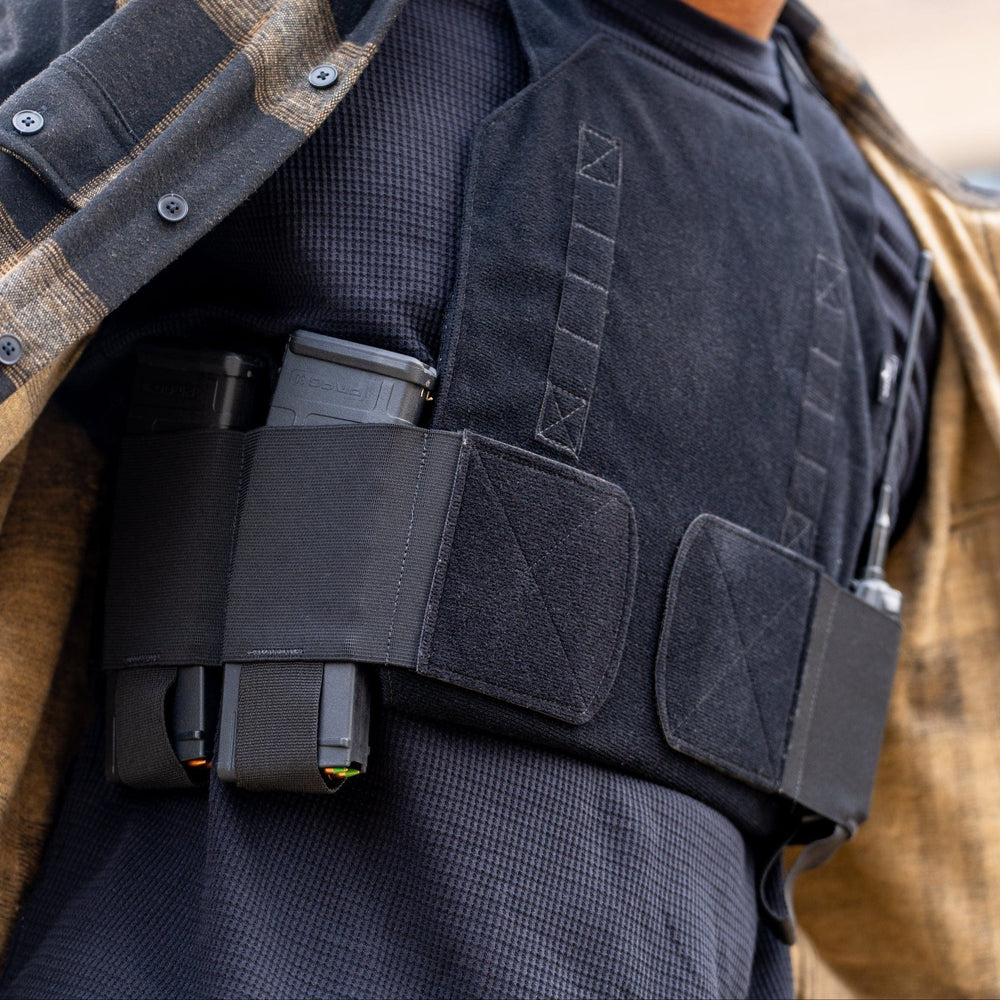 A person in a flannel shirt subtly showcases the Discreet Defender Bundle - Agile PE Armor by Premier Body Armor beneath. The vest, with two magazines in its front elastic pouches, offers concealed protection, while a blurred building adds mystery to the scene.