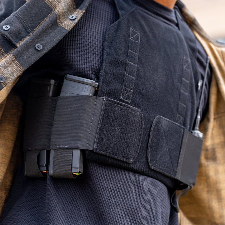 A person in a flannel shirt subtly showcases the Discreet Defender Bundle - Agile PE Armor by Premier Body Armor beneath. The vest, with two magazines in its front elastic pouches, offers concealed protection, while a blurred building adds mystery to the scene.