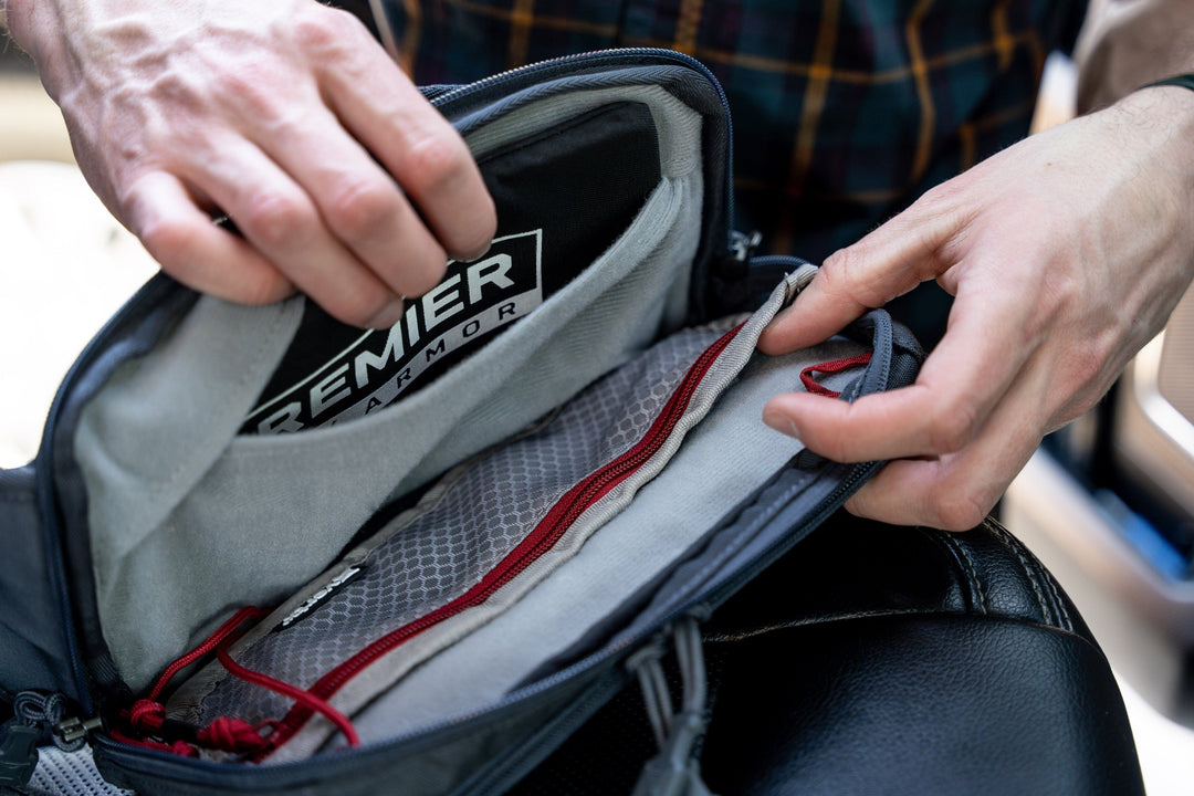 A person opens a backpack pocket to show a Premier Armor ballistic panel inside the compartment, with the person's hands holding the fabric apart and a plaid shirt partially visible in the background.