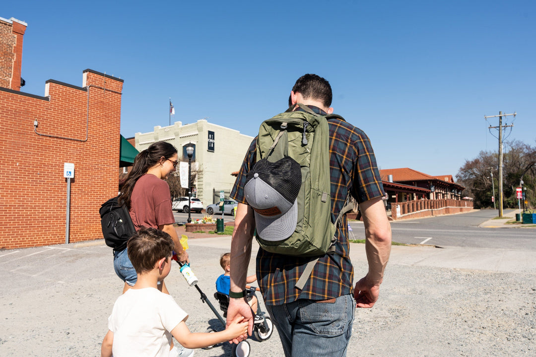 A man with a green backpack and baseball cap walks outdoors, holding a child's hand. Two children and a woman with a black backpack walk alongside him on a sunny day, with brick buildings visible in the background.