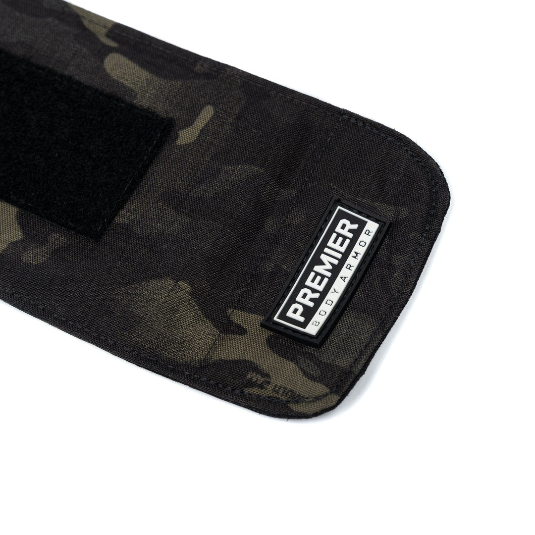 Close-up of a black and green camouflage fabric made from ballistic material, featuring a "PREMIER BODY ARMOR" label stitched in white and black on the bottom right corner. A small section of black hook-and-loop fastener is visible on the left side of the fabric, part of Premier Body Armorโs Bulletproof Can Cooler.