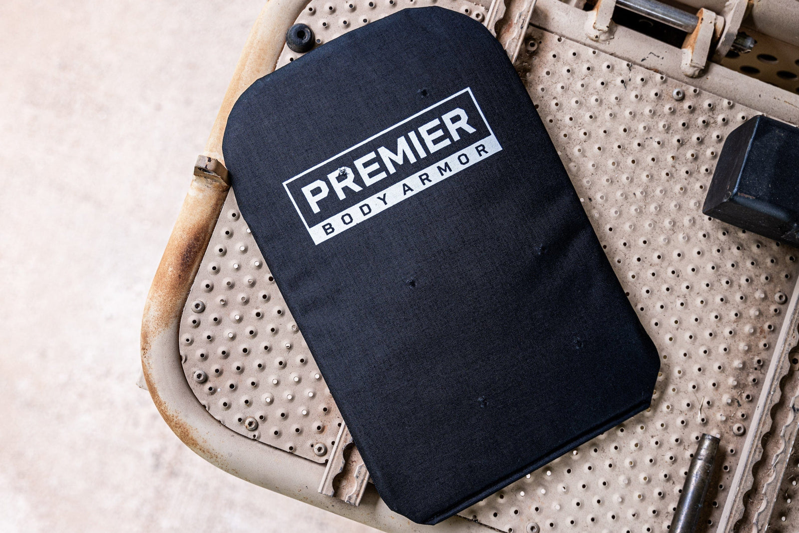 A black rectangular ballistic plate labeled "Premier Body Armor" rests on a beige perforated metal surface.