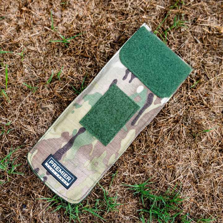 A camouflage-patterned Bulletproof Can Cooler by Premier Body Armor, with a green Velcro flap and patch, lies on dry grass. Infused with ballistic fibers, it features a "PREMIER" label for durability and protection.