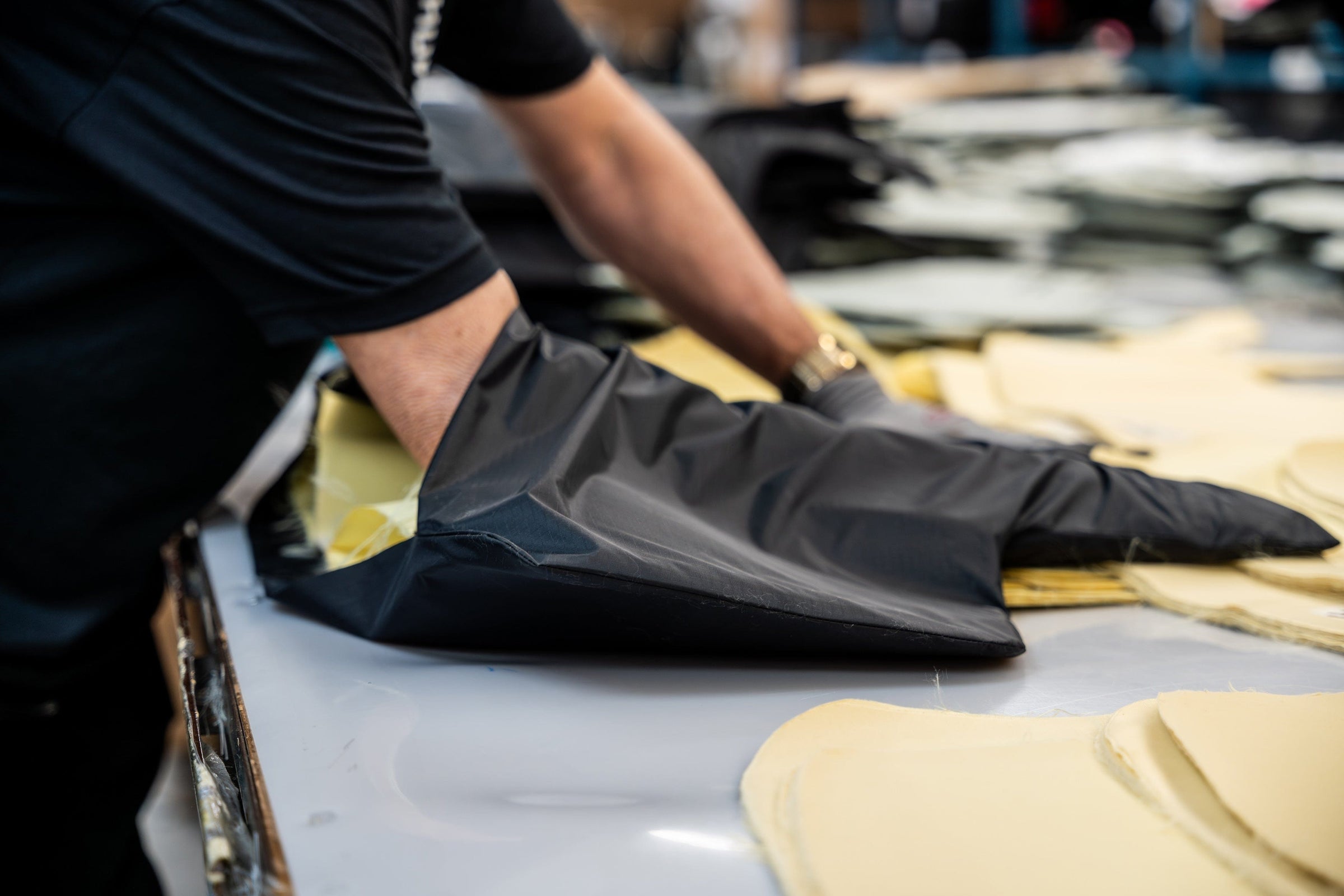 A person wearing a black shirt and long black gloves is handling large sheets of yellow foam or fabric on a worktable. More similar sheets are spread out across the surface.