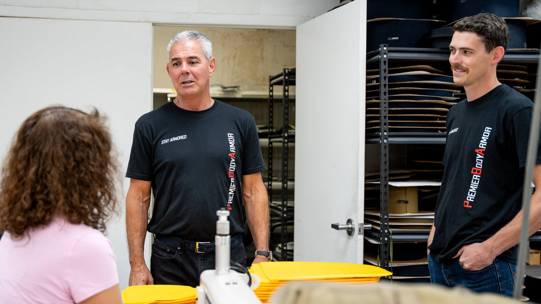 Two men in black "Premier Body Armor" shirts stand and talk to a seated woman in a workspace with yellow bins and shelves holding large flat objects. The setting appears to be a workshop or storage room.