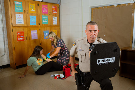 A police officer holding a ballistic shield stands in a classroom while a woman in medical gloves attends to a seated person near a first aid kit; colorful posters are on the wall in the background.