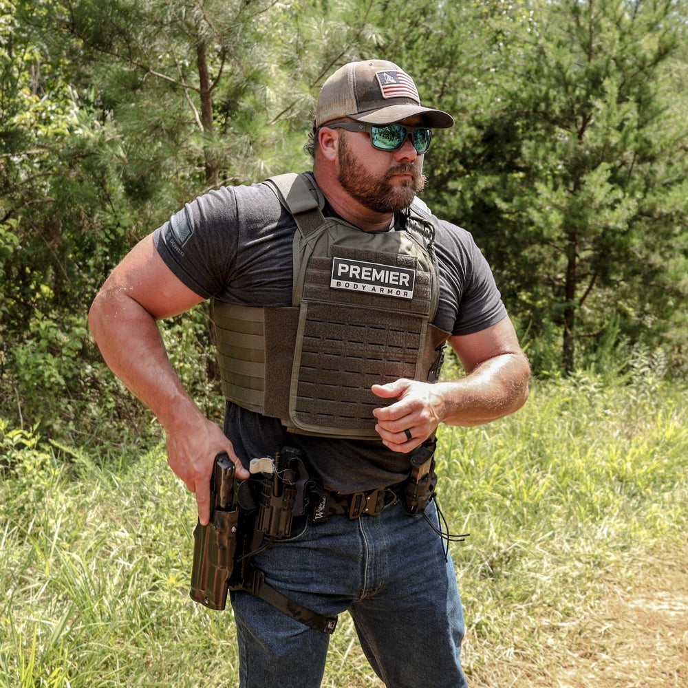 A man stands outdoors in a wooded area, dressed in a dark T-shirt, blue jeans, and a baseball cap. Wearing sunglasses and the Premier Body Armor Core Mission Plate Carrier with Level IIIA Cummerbund Armor featuring MOLLE webbing, he has a holstered firearm at his waist. Trees and grass form the background.