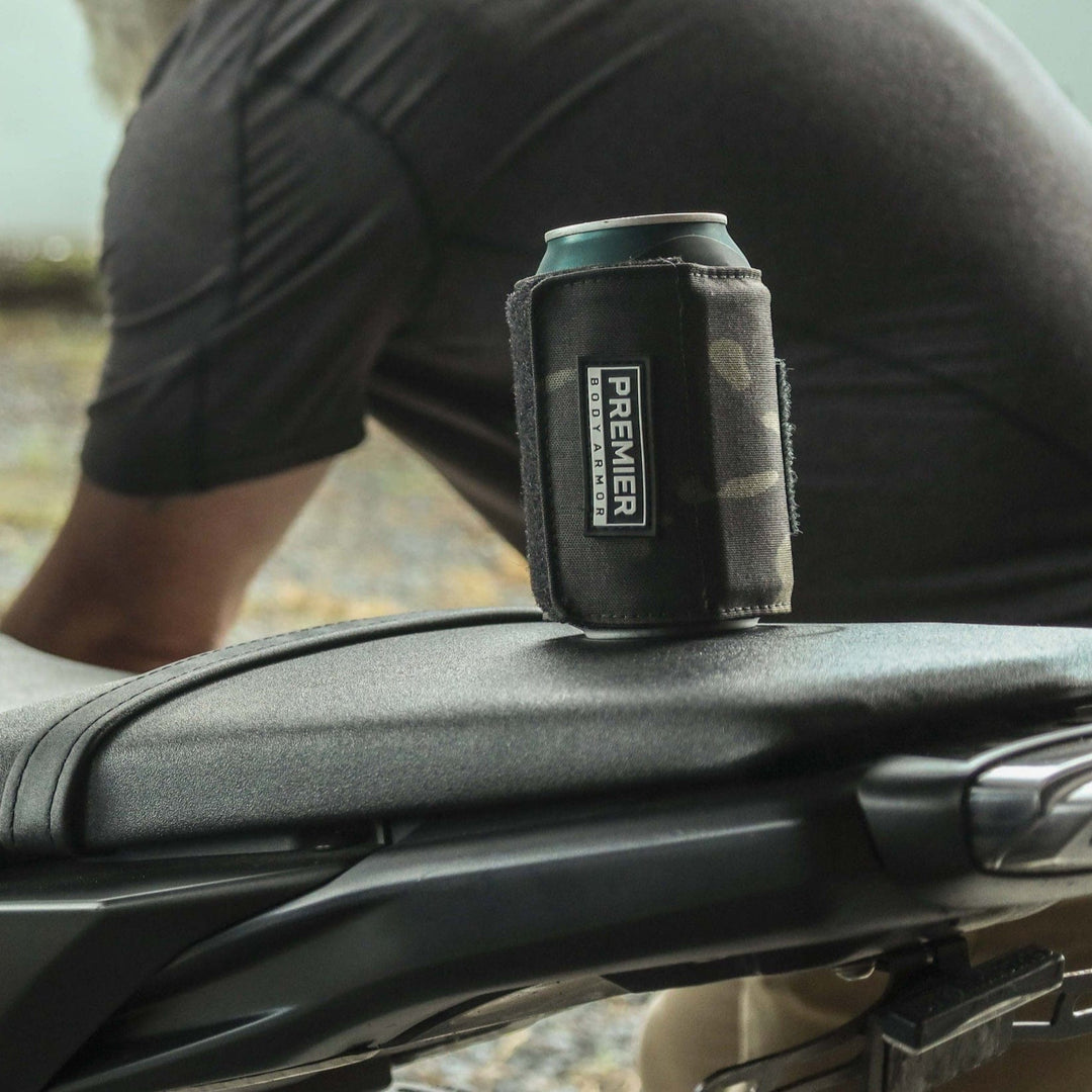 On a motorcycle seat sits a can wrapped in a black Premier Body Armor koozie. The Bulletproof Can Cooler, made from ballistic material, ensures your drink stays secure. In the background, a person in a dark shirt is partially visible, leaning forward against an outdoor backdrop.