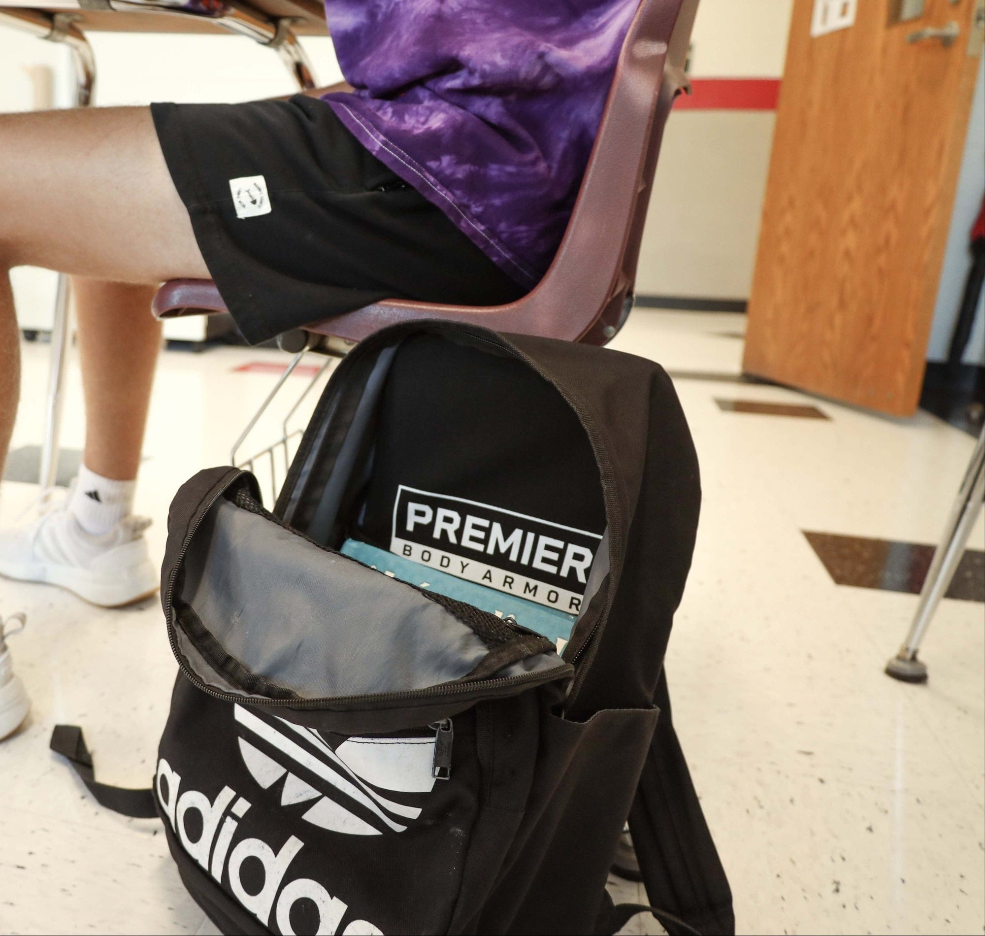 A student wearing black shorts sits at a desk. On the floor is a black Adidas backpack with a visible Premier Body Armor panel and a book inside. The classroom has tiled floors and a wooden door in the background.