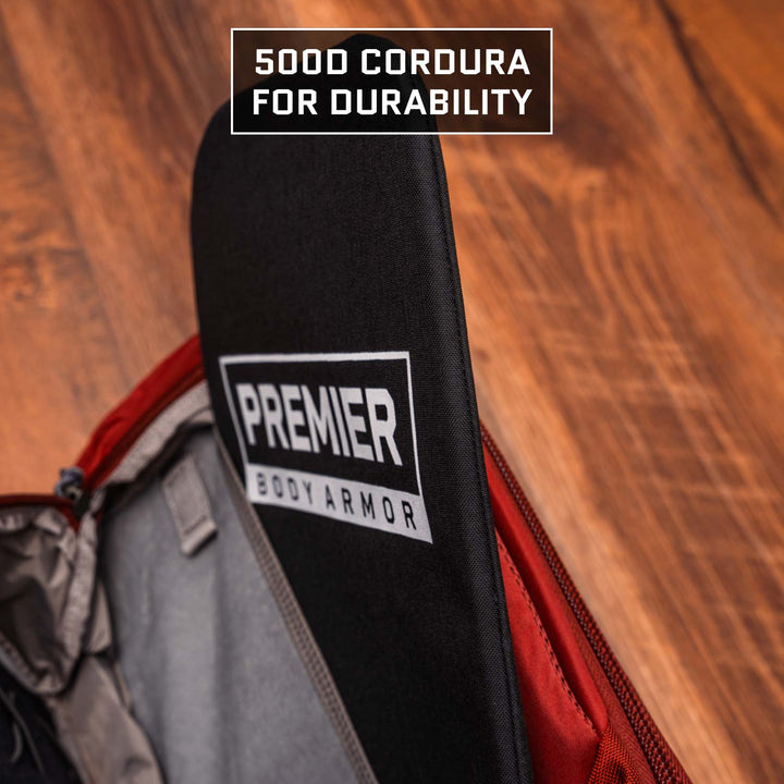 A Multicam Black 9 x 13.5 Level IIIA Armor Insert by Premier Body Armor is shown inside a red backpack on a wooden floor. "500D Cordura for durability" appears above.