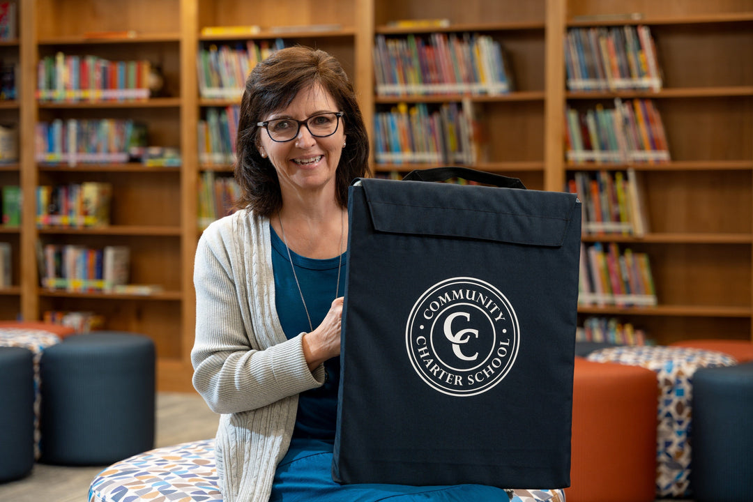 A woman with glasses and shoulder-length hair sits in a library, smiling and holding a black bag with the "Community Charter School" logo; bookshelves filled with books are in the background.