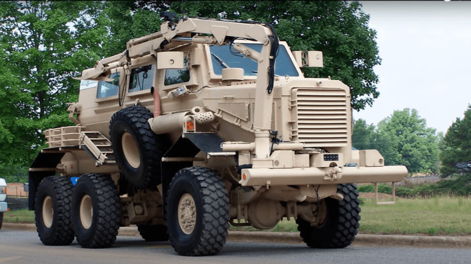 A tan, heavily armored military vehicle with large tires and external equipment is parked on a paved surface, surrounded by greenery and trees in the background.