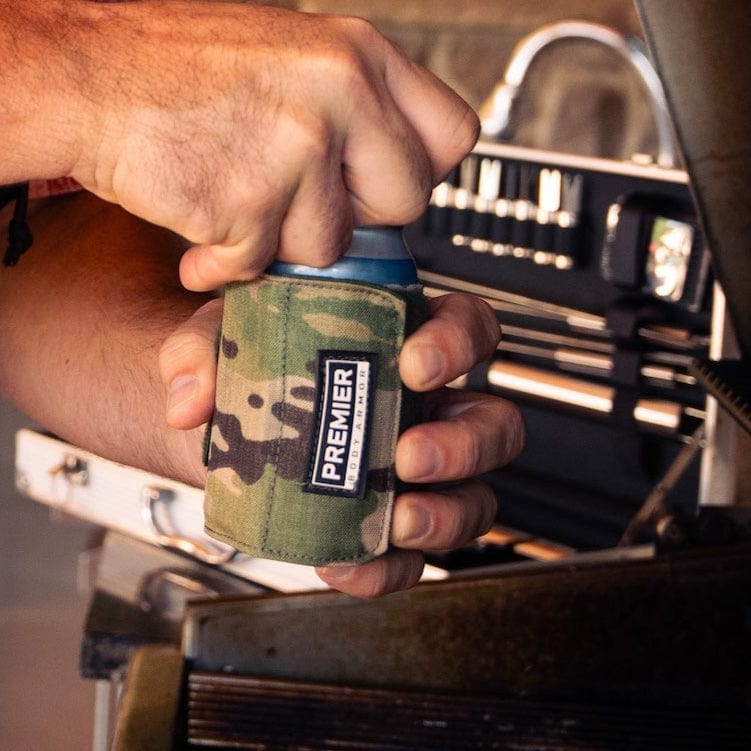 A hand holds a "Premier Body Armor" Bulletproof Can Cooler wrapped in camouflaged ballistic fibers around a can, while the person stands near a barbecue grill with visible knobs and cooking surface in the background.