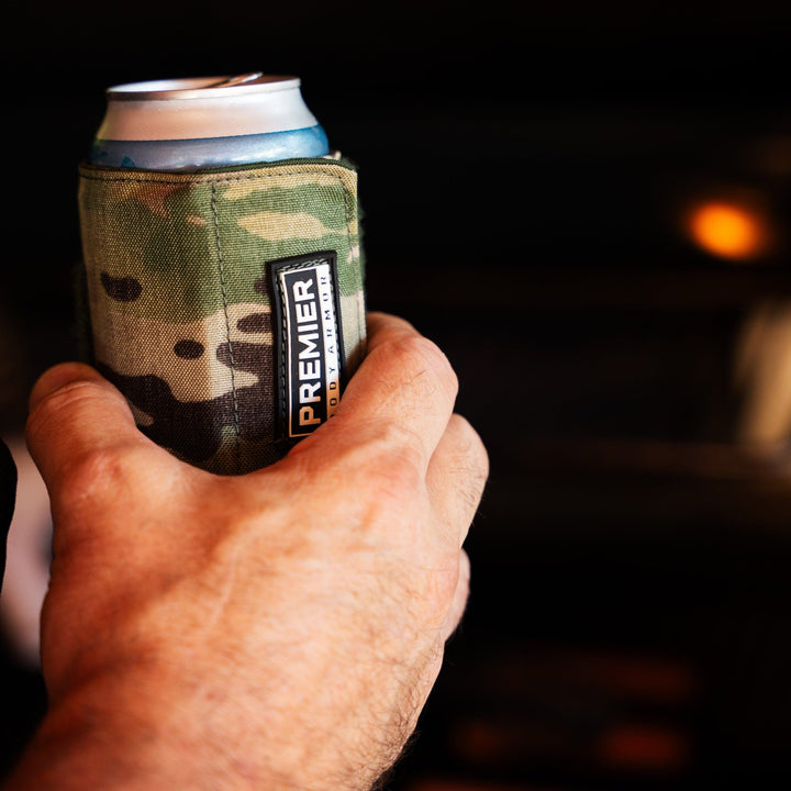 A hand holds a Premier Body Armor Bulletproof Can Cooler with a camouflage pattern and the word "PREMIER" visible on the side, against a dark, blurred background highlighting the innovative cooler made from ballistic fibers.