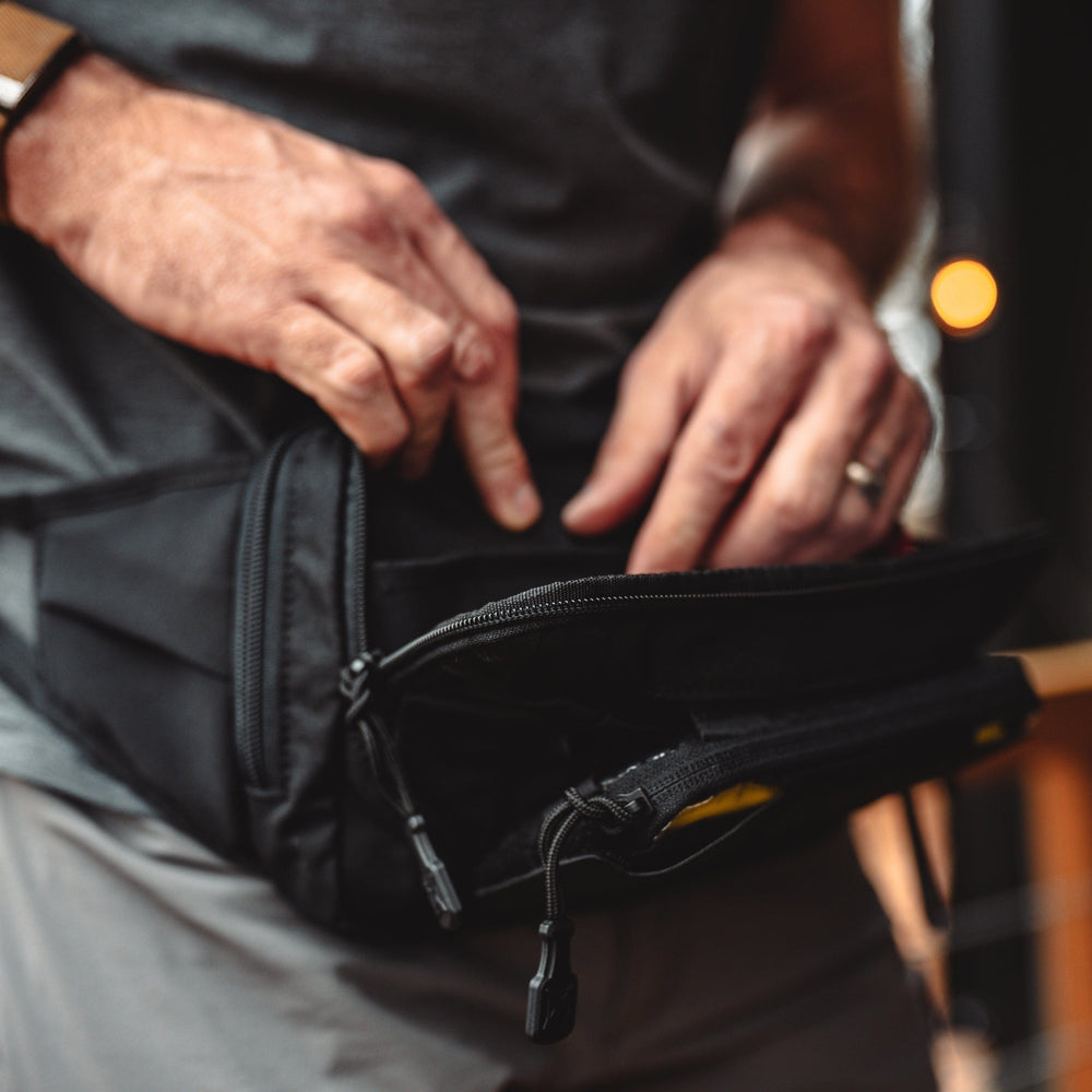 A person wearing a gray shirt and beige pants is shown opening the zipper of a black Vertx SOCP Tactical Fanny Pack with precision similar to tactical utility gear.