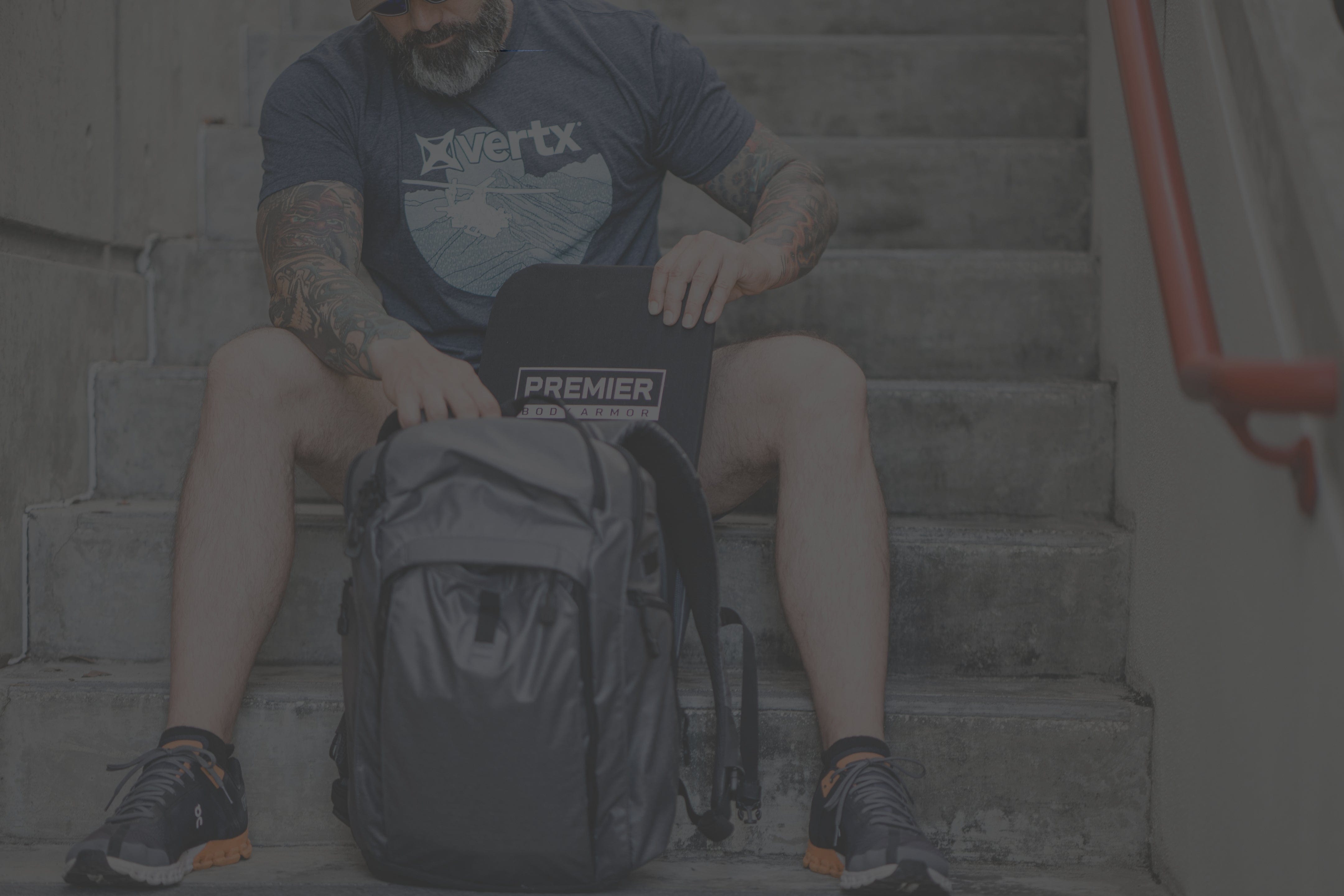 A bearded man with tattoos sits on outdoor steps, placing a "Premier Body Armor" panel into a black backpack. He is wearing a blue "Vertx" t-shirt, black shorts, and sneakers.