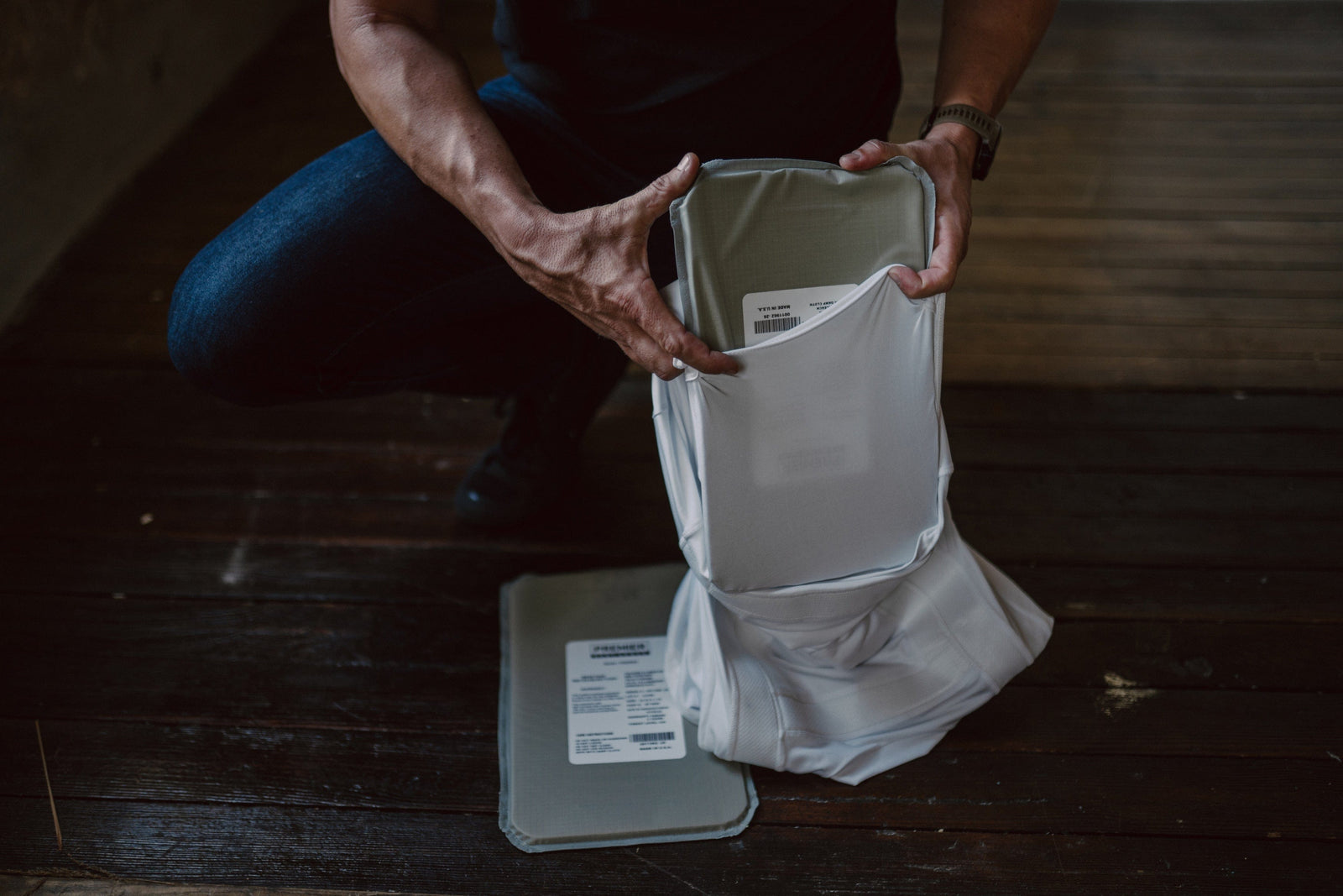 A person kneels on a wooden floor, holding and inspecting a white, rectangular fabric pouch with labels. The pouch's open top reveals its interior, while its flat lid and additional labels rest on the floor nearby.