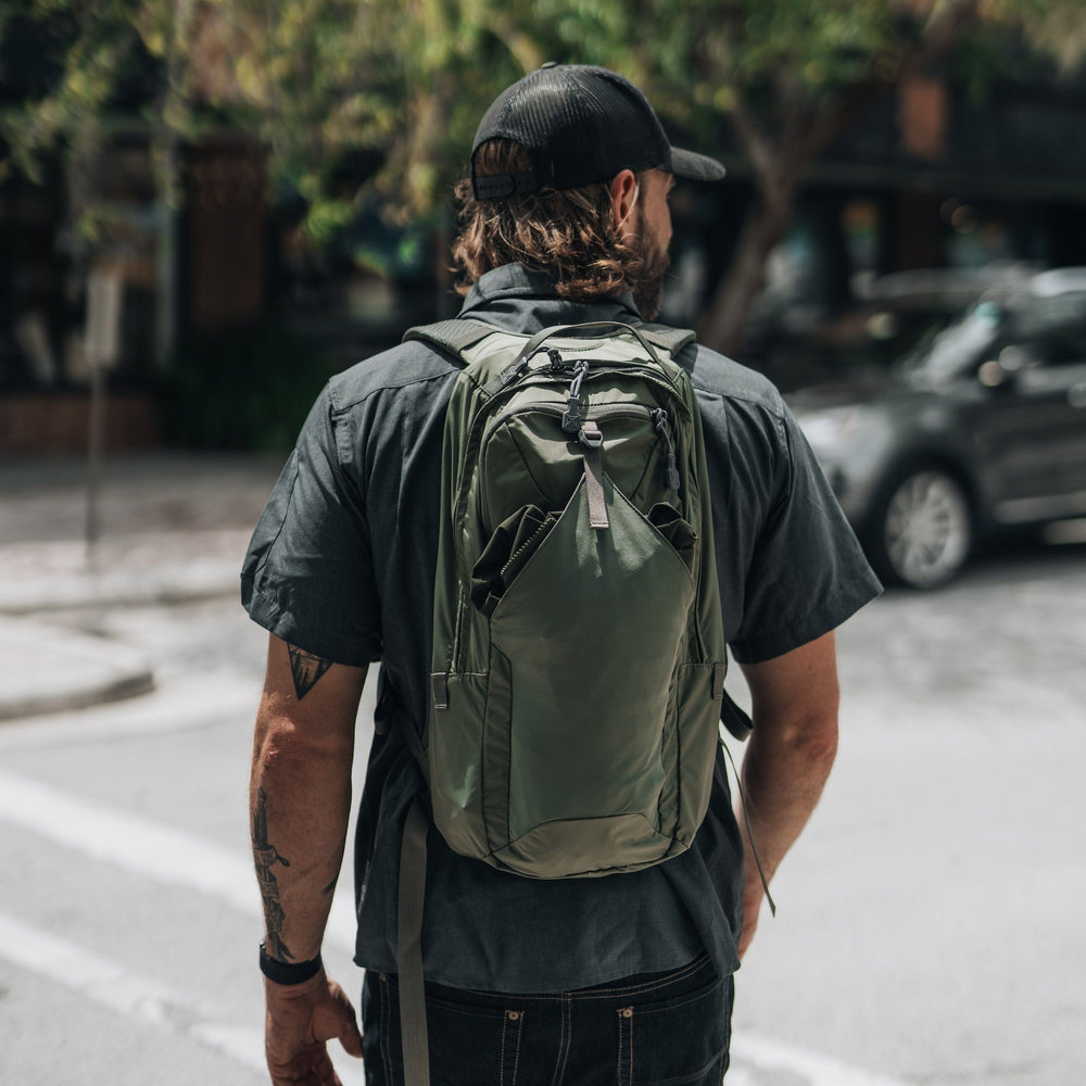 A man wearing a black shirt, jeans, and cap stands on a city street corner with a tattoo on his left forearm. He carries the Vertx Long Walks Pack 15L backpack in green, with cars and trees visible in the background.