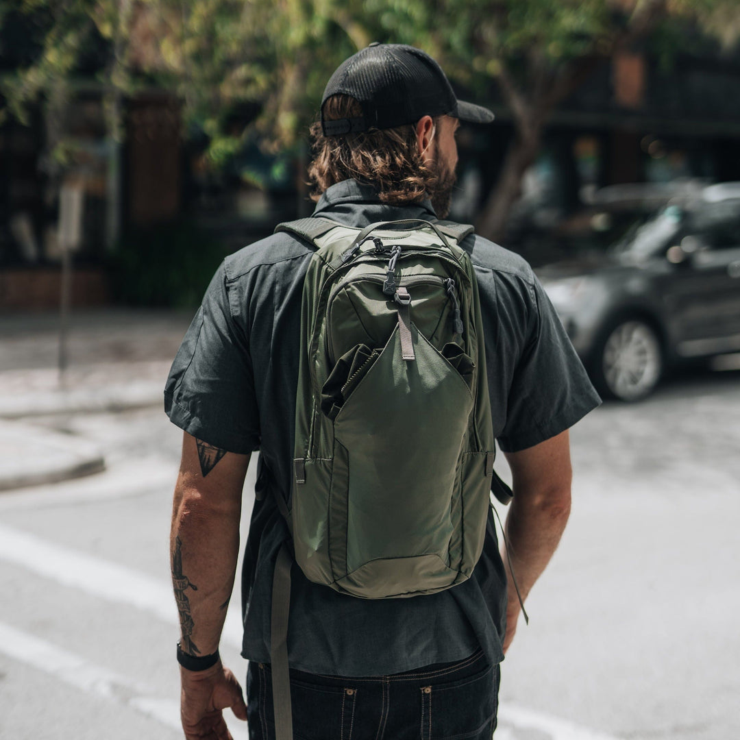 A man wearing a black shirt, jeans, and cap stands on a city street corner with a tattoo on his left forearm. He carries the Vertx Long Walks Pack 15L backpack in green, with cars and trees visible in the background.