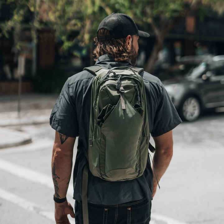 A man wearing a black shirt, jeans, and cap stands on a city street corner with a tattoo on his left forearm. He carries the Vertx Long Walks Pack 15L backpack in green, with cars and trees visible in the background.