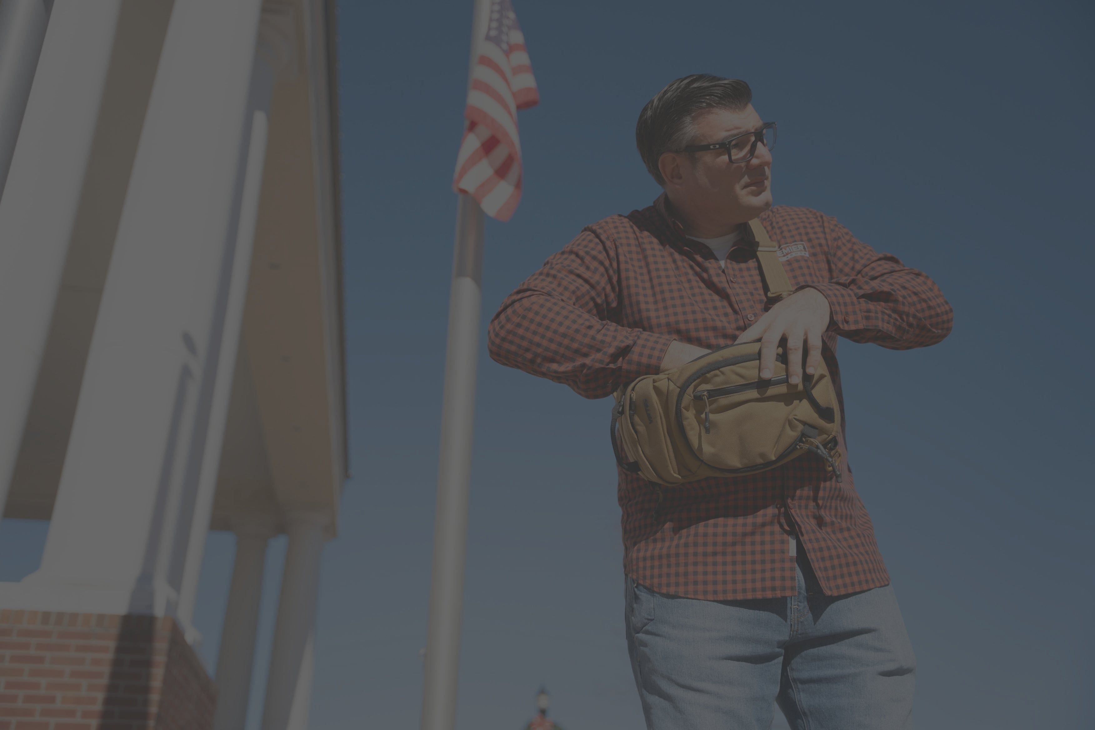 A man wearing sunglasses and a plaid shirt stands outdoors next to a building with columns and an American flag, reaching into a tan shoulder bag. The sky is clear and blue.