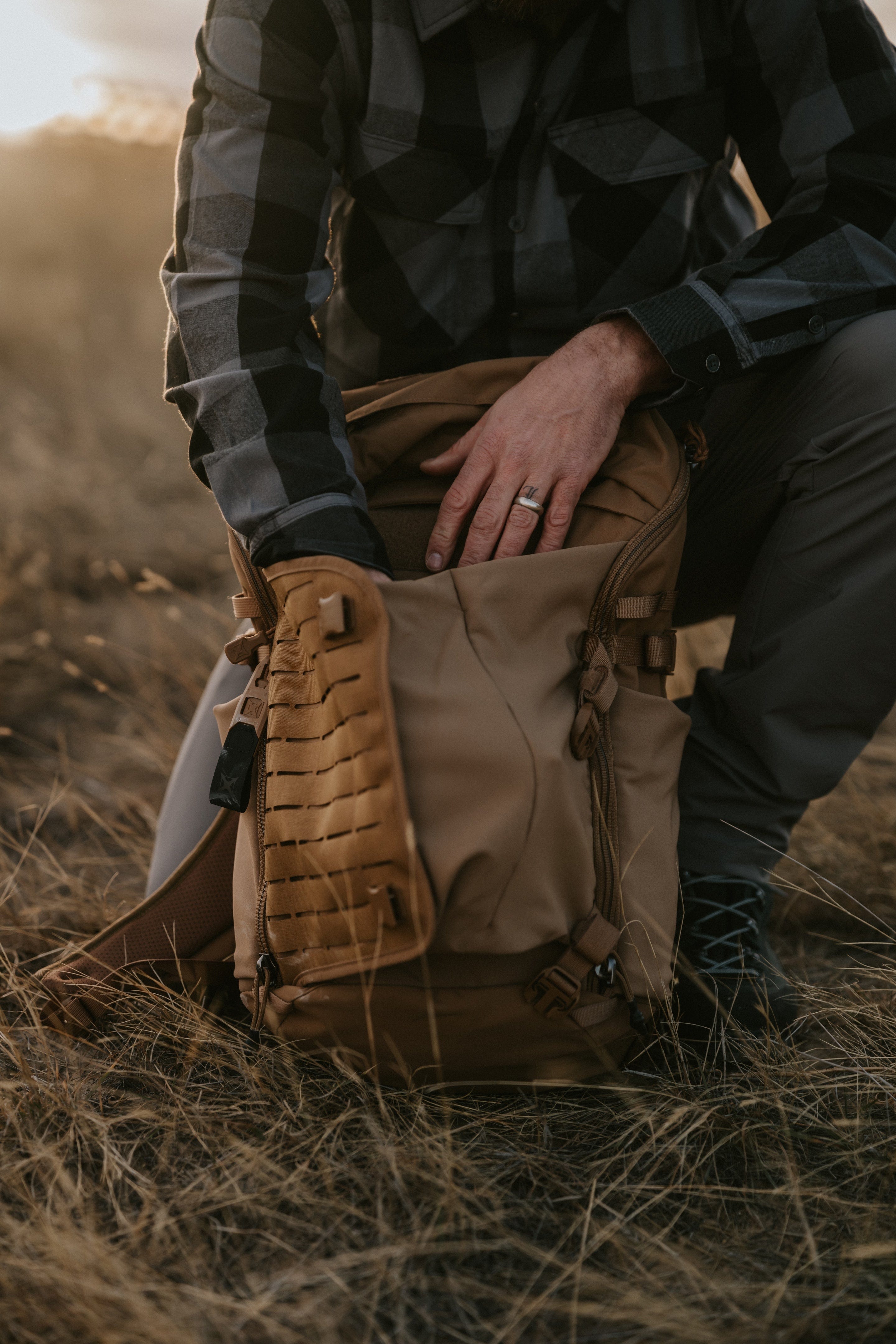 A person wearing a plaid shirt and gray pants kneels on dry grass while opening a tan backpack, with one hand reaching inside the bag.