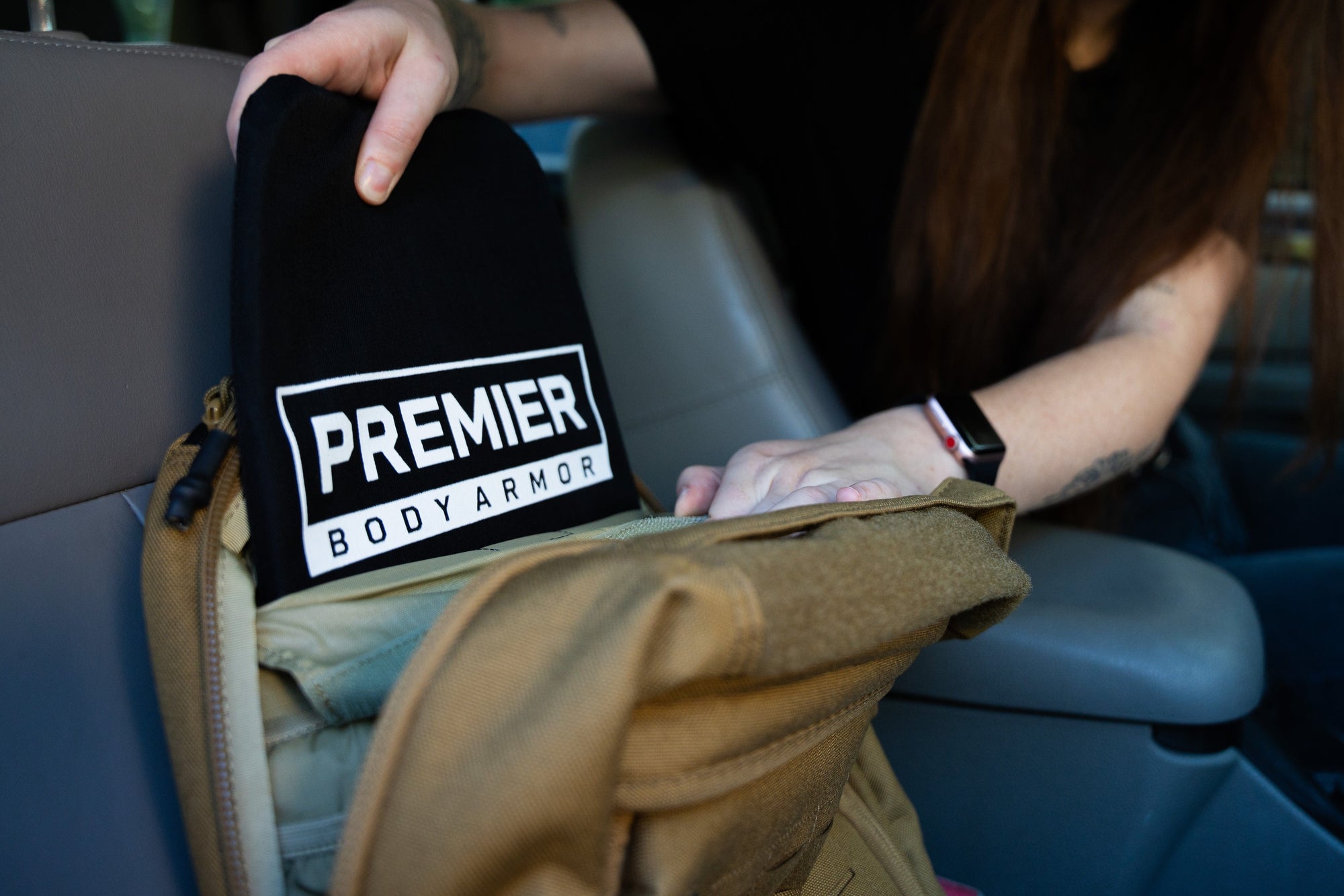 A person places a black object labeled "Premier Body Armor" into the front pocket of a tan backpack while seated in a vehicle.