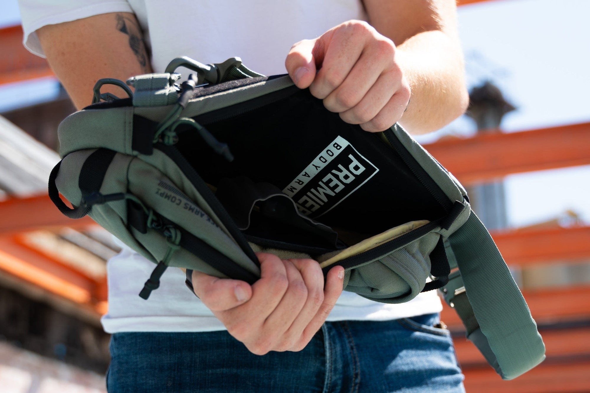 A person wearing a white shirt and jeans holds open a gray crossbody bag, revealing a “PREMIER BODY ARMOR” label inside. The background is outdoors with orange metal beams visible.