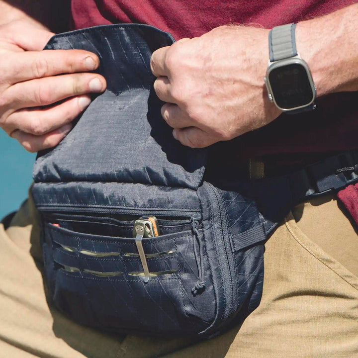 A person in a red shirt and beige pants adjusts their Viktos Upscale Hip Pack, showcasing its tactical design. Their left wrist bears a smartwatch. This black waist pack by Viktos/Premier offers multiple compartments, CCW access, and a clip with a small tool for convenience.