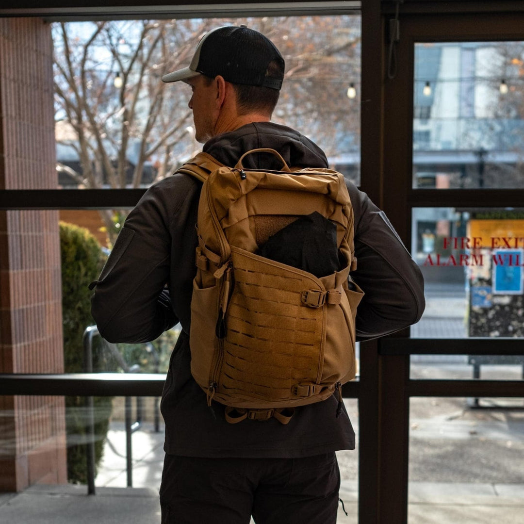 A person wearing a baseball cap and dark jacket stands indoors with their back to the camera, carrying a Vertx Siege 25L Tactical Pack featuring versatile functionality and multiple compartments. Through the glass door, trees and a sidewalk are visible outside.