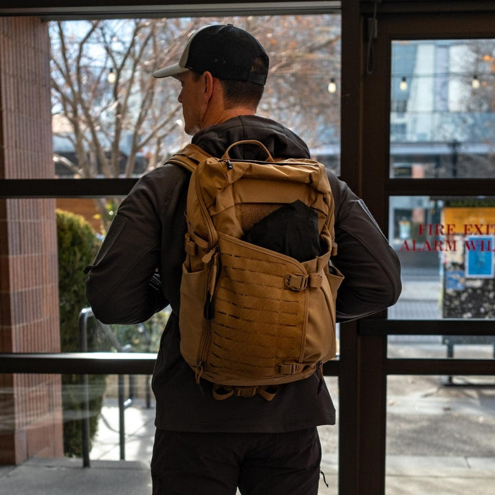 A person wearing a baseball cap and dark jacket stands indoors with their back to the camera, carrying a Vertx Siege 25L Tactical Pack featuring versatile functionality and multiple compartments. Through the glass door, trees and a sidewalk are visible outside.