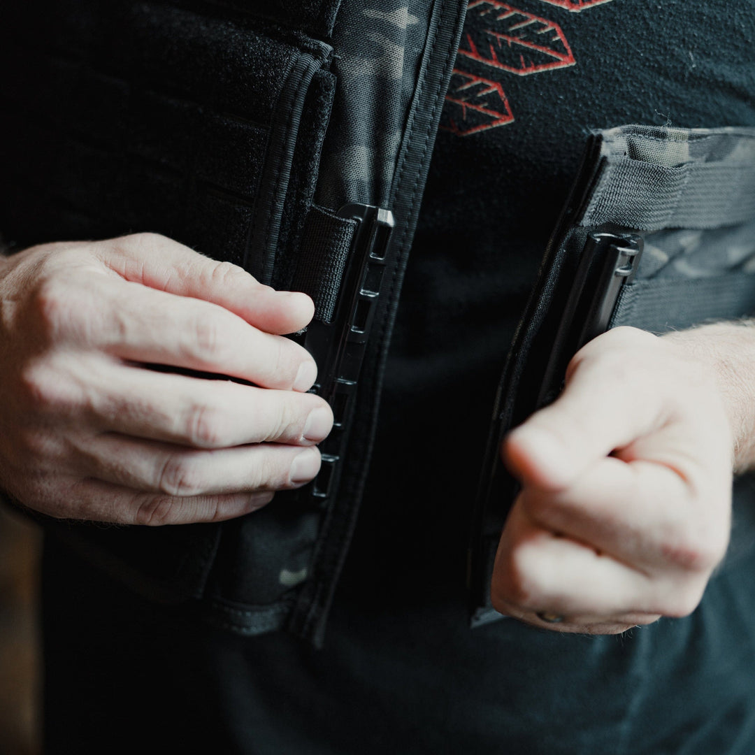 A person adjusts the straps of a black Core Mission Plate Carrier by Premier Body Armor, featuring MOLLE webbing for gear attachment, over a black shirt with a partially visible red feather pattern. The focus is on their hands and vest against a softly blurred background.