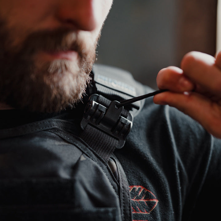 A bearded person adjusts the strap of a black Premier Body Armor Core Mission Plate Carrier with Level IIIA Cummerbund Armor. The focus is on the shoulder clasp, highlighting its high-speed performance, as they wear a black shirt with subtle molle webbing and a red logo in the backdrop.