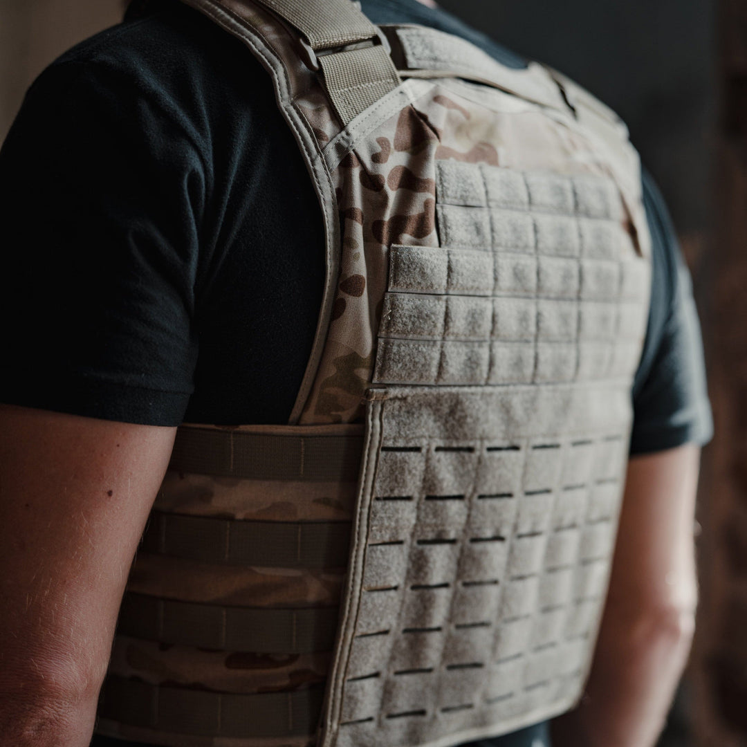 A man wears a Premier Body Armor Core Mission Plate Carrier w/ Level IIIA Cummerbund Armor over a black t-shirt. The vest, built for high-speed performance, features traditional molle webbing, padded protection, and multiple horizontal straps. He stands indoors against a dark background.