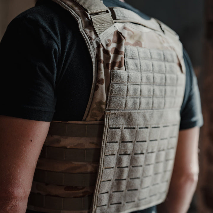 A man wears a Premier Body Armor Core Mission Plate Carrier w/ Level IIIA Cummerbund Armor over a black t-shirt. The vest, built for high-speed performance, features traditional molle webbing, padded protection, and multiple horizontal straps. He stands indoors against a dark background.