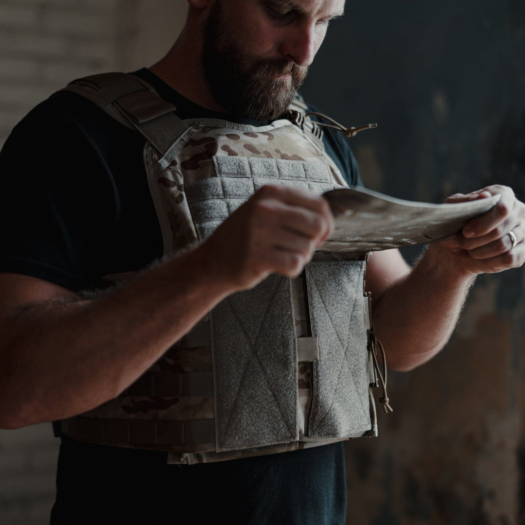 In a dimly lit room with textured walls, a bearded man examines a piece of fabric on his Core Mission Plate Carrier w/ Level IIIA Cummerbund Armor by Premier Body Armor. The tactical vest, equipped with MOLLE webbing, appears tailored for his core mission needs.