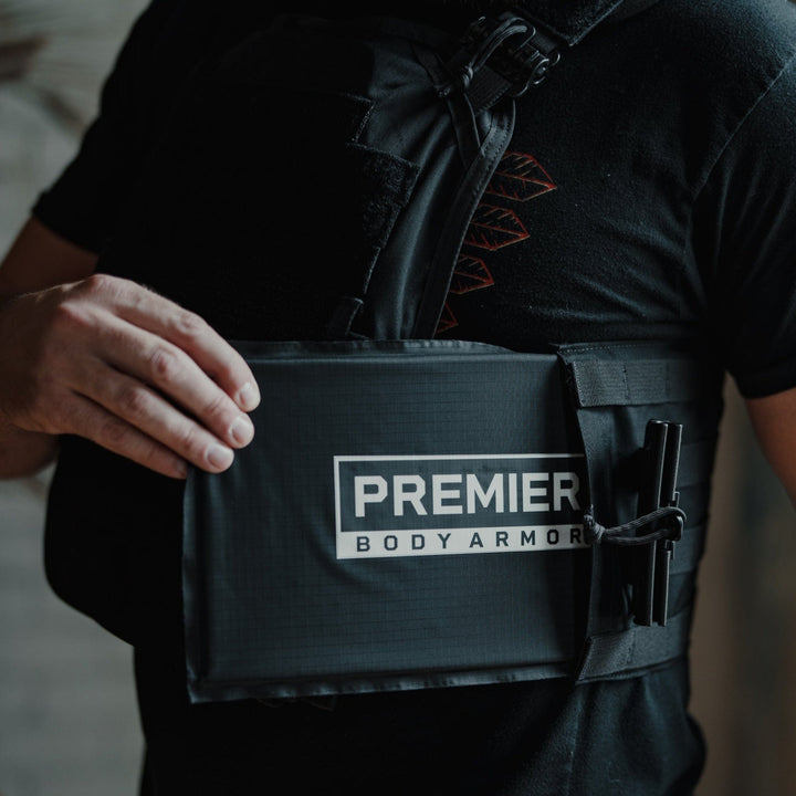 A bearded man in a black shirt and jeans adjusts his Core Mission Plate Carrier w/ Level IIIA Cummerbund Armor, branded "Premier Body Armor." The image emphasizes the armor's front panel and label, showcasing MOLLE webbing details against a softly blurred background.
