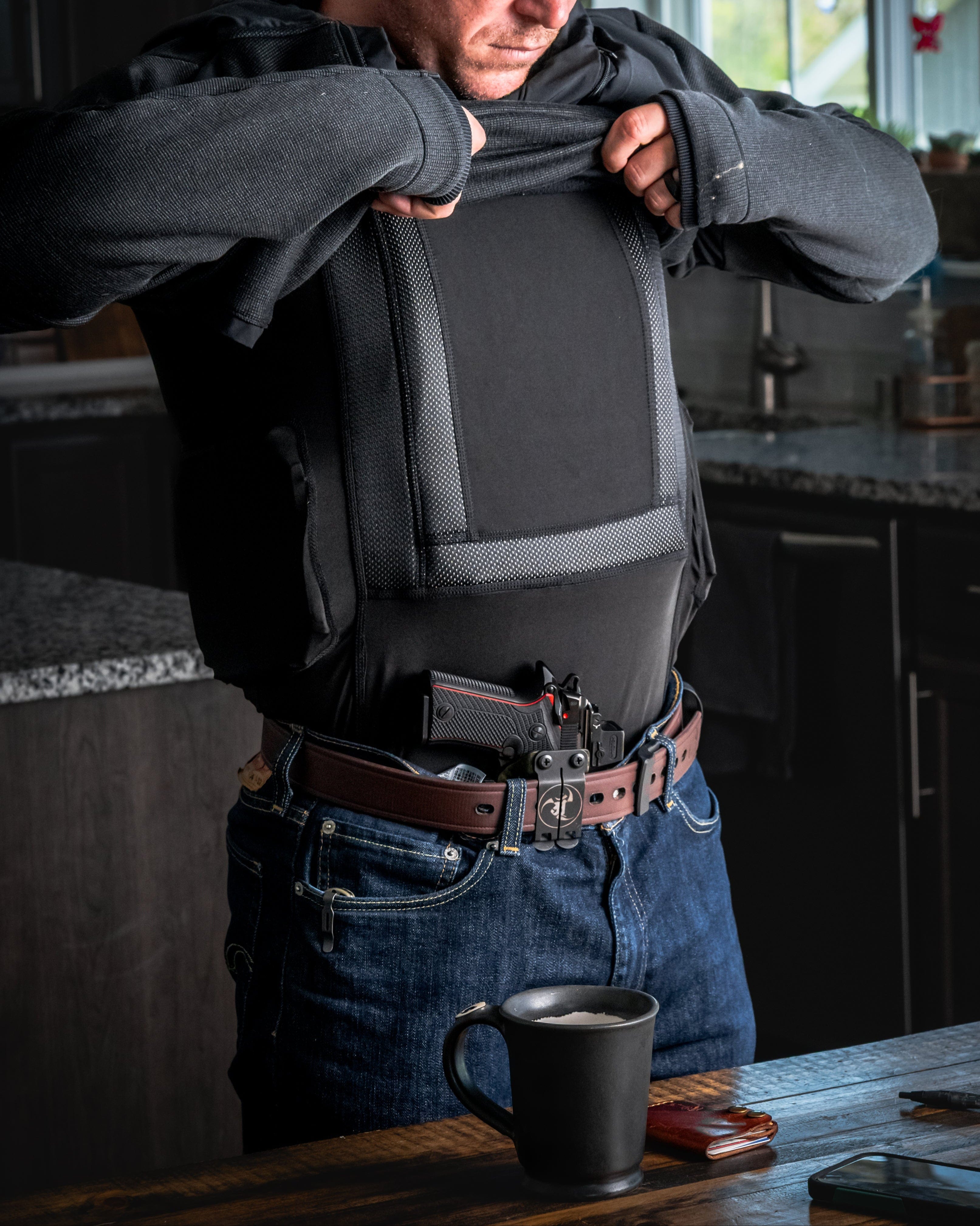A person lifts their shirt to reveal a black bulletproof vest and a holstered handgun on their belt. They are standing in a kitchen, with a mug, wallet, and phone on a nearby counter.