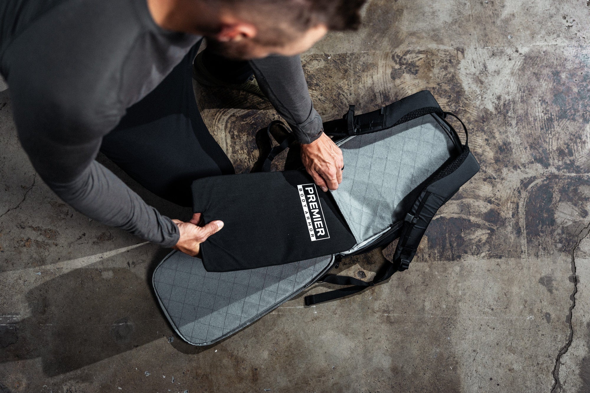 A person places a Premier body armor panel into the back compartment of a black backpack on a concrete floor.