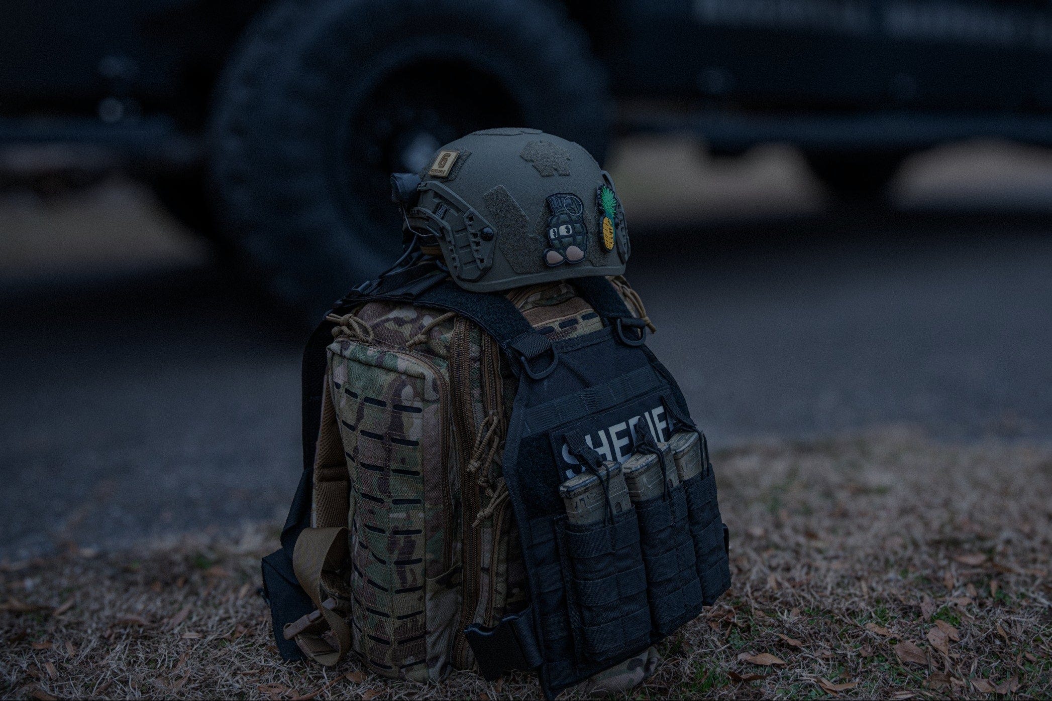 A tactical vest labeled "SHEIRFF" with attached pouches and gear sits on the ground next to a military-style backpack and helmet in front of a large, dark vehicle.