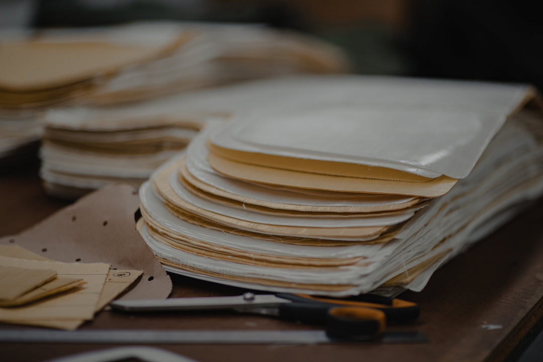 A stack of rectangular, layered fabric or paper pieces with rounded edges sits on a table, next to a pair of scissors and more stacked materials in the background.