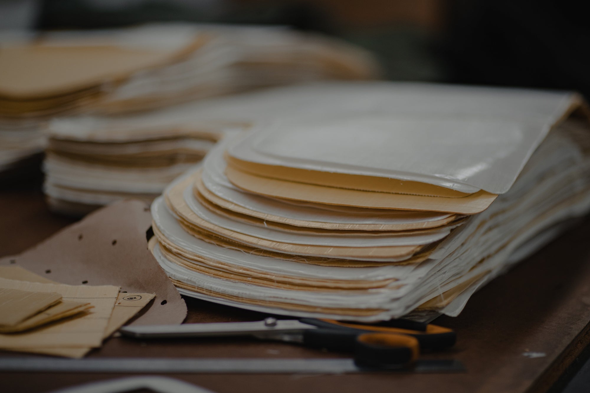 A stack of rectangular, layered fabric or paper pieces with rounded edges sits on a table, next to a pair of scissors and more stacked materials in the background.