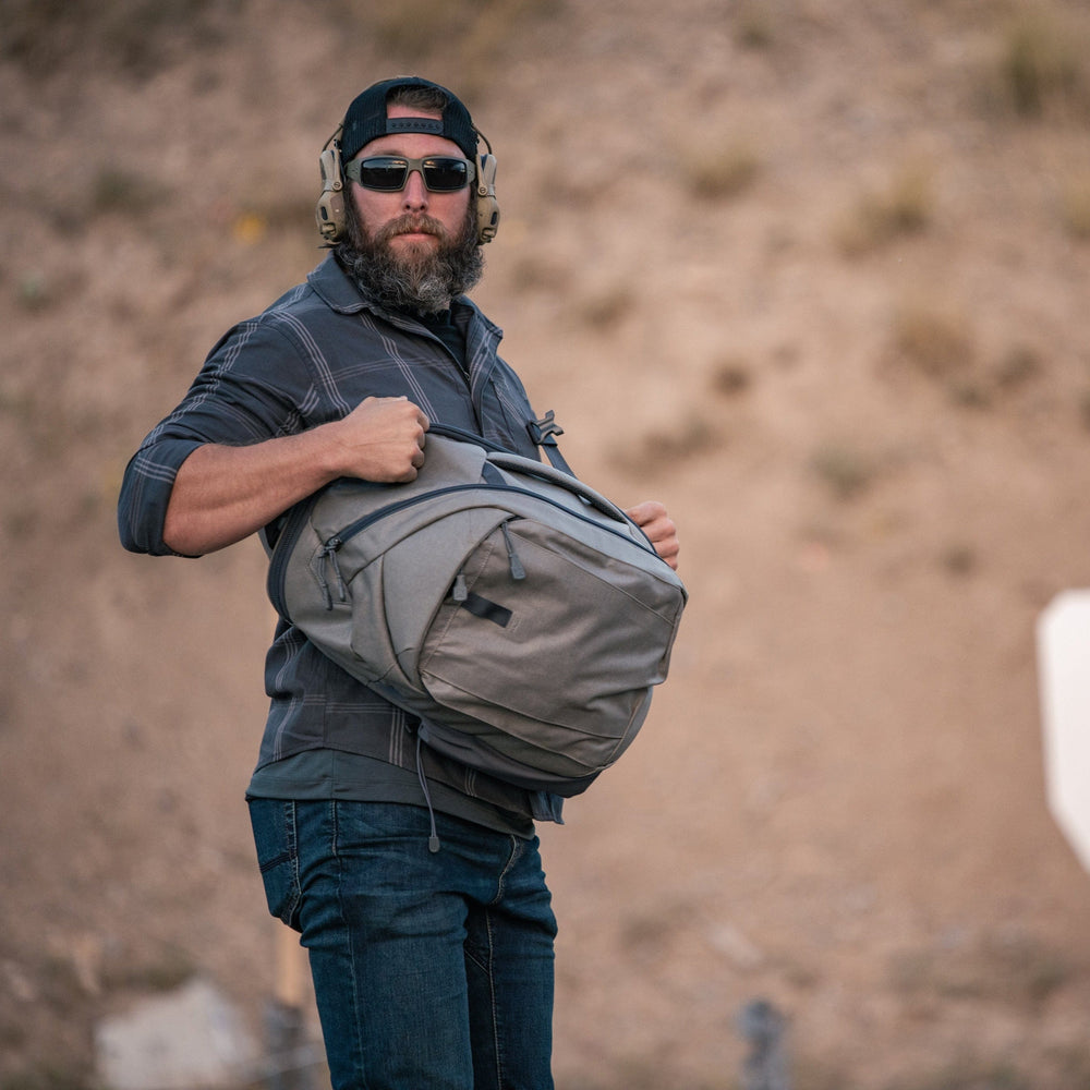 A man outdoors with a dirt backdrop wears sunglasses, a hat, and headphones while carrying a gray Vertx Commuter 3.0 Sling with body armor insert. Metal stands are on the ground and a white target is clearly visible in the background.