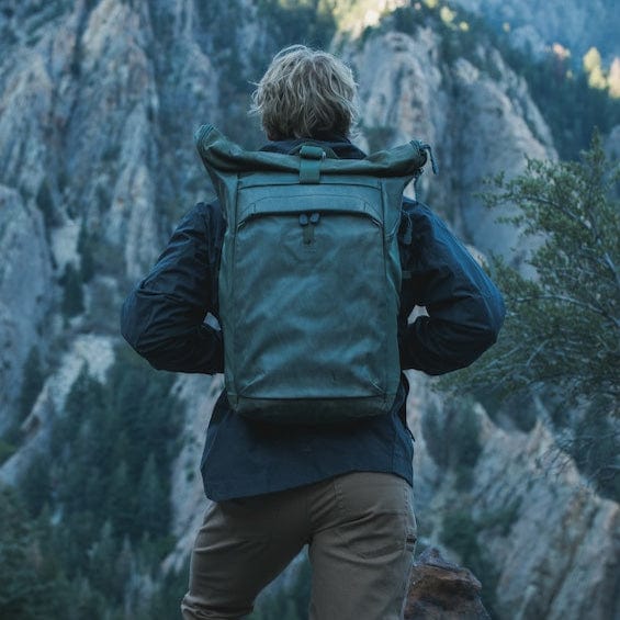 A person with light hair stands with their back to the camera, wearing a Vertx Ruck Roll Pack and outdoor clothing. They gaze at a mountainous landscape filled with trees and rocky cliffs, ready for adventure.